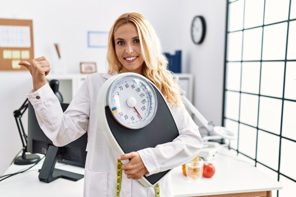 beautiful blonde nutritionist woman holding weight machine to balance weight loss pointing thumb up to the side smiling happy with open mouth